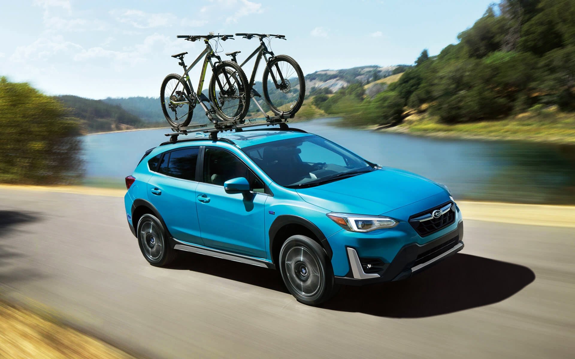 A blue Crosstrek Hybrid with two bicycles on its roof rack driving beside a river | Burke Subaru in Cape May Court House NJ